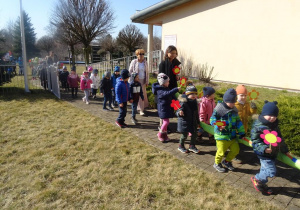 Dzieci wraz z panią dyrektor maszerują z wiosennymi plakatami, panią Wiosną pod budynkiem przedszkola.